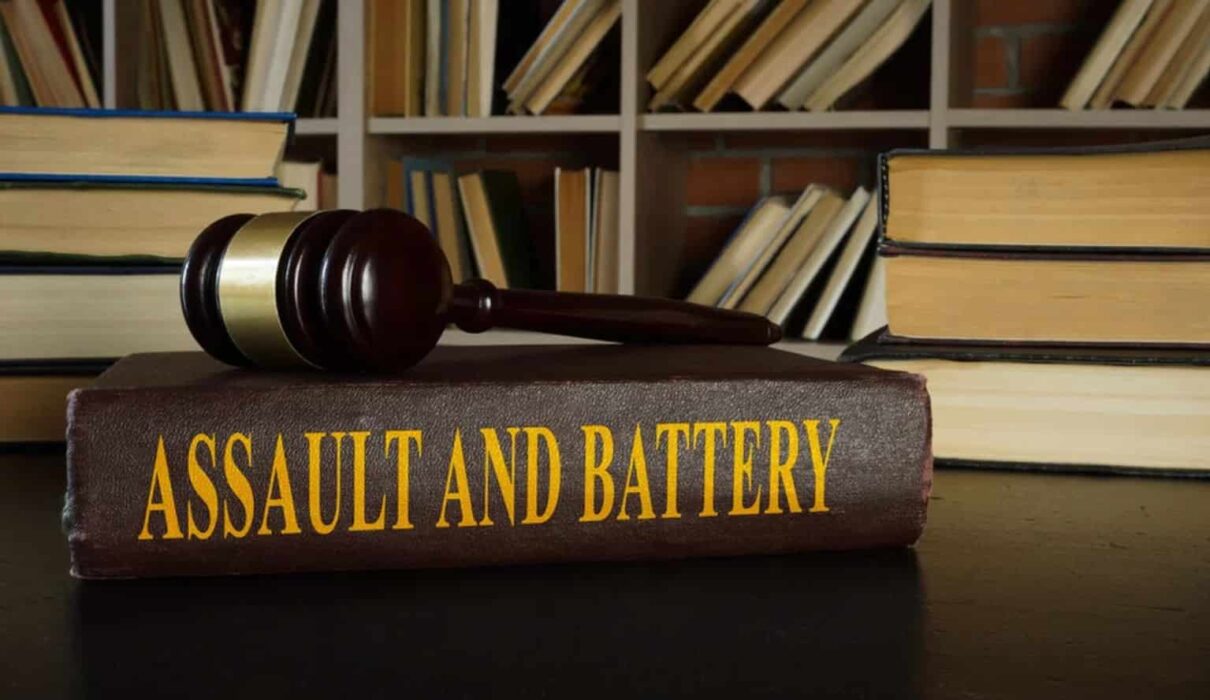 A courtroom-themed illustration representing Assault and Battery, showing a victim with visible injuries speaking to a lawyer while legal documents, a gavel, and scales of justice symbolize a civil and criminal legal case.